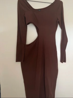Brun långärmad klänning med cut-out - Trendig brun klänning med lång ärm och cool cut-out detalj i midjan. Klänningen har en kroppsnära passform och är tillverkad i ett stretchigt material som ger en snygg siluett. Perfekt för dig som vill sticka ut med en unik look.