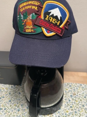 Mörkblå Dsquared2 keps med patches - Snygg mörkblå keps från Dsquared2 med två stora broderade patches framtill. Ena patchen har texten 'Campfire 1964' och motiv av eld och skog, den andra har bergsmotiv och texten 'Caten Cadore 1964'. Justerbar baktill och klassisk böjd skärm.