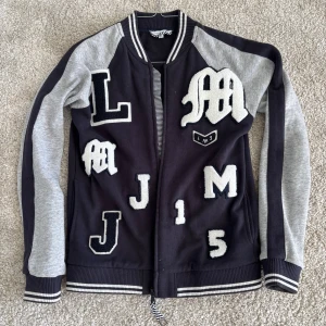 Mörkblå och grå bomberjacka Little Marc Jacobs - Snygg jacka/kofta från Little Marc Jacobs i mörkblått och grått med snygga stora applikationer/ bokstäver på framsidan. Jackan har ribbade muddar med ränder och dragkedja framtill. Rök-/djurfritt hem.