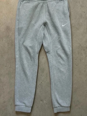 Gråa Nike mjukisbyxor - Snygga grå mjukisbyxor från Nike med klassisk vit logga på vänster lår. Byxorna har resår i midjan, muddar vid bensluten och sidofickor samt en bakficka. Tillverkade i mjukt sweatshirtmaterial som är perfekt för chill dagar.