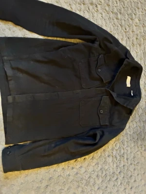 Svart overshirt med bröstfickor från Jack and Jones - Stilren svart overshirt med två stora bröstfickor med lock och knappstängning. Klassisk krage och knappar framtill. Perfekt lager-på-lager-plagg i ett mjukt tyg som ger en avslappnad look.