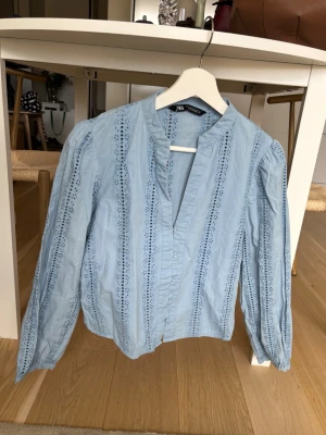 Ljusblå blus med broderade detaljer Zara - Supersöt ljusblå blus från Zara med broderade hålband längs hela plagget. Blusen har v-ringning utan krage, knäppning framtill och långa puffiga ärmar. Perfekt till jeans eller kjol för en fräsch look. Materialet är bomull som känns lätt och skönt.