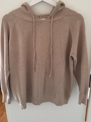 Beige stickad hoodie med huva - En stilren beige stickad hoodie med huva och dragsko. Tröjan har långa ärmar och ribbade muddar vid ärmslut och nederkant. Perfekt nu inför sommaren.