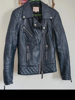 Svart skinnjacka imitation  från Livelo - Snygg svart skinnjacka från Livelo med coola quiltade detaljer på axlar och nederkant. Jackan har flera dragkedjor framtill, snedställd dragkedja och bred krage. Perfekt för dig som vill ha en edgy och trendig look.