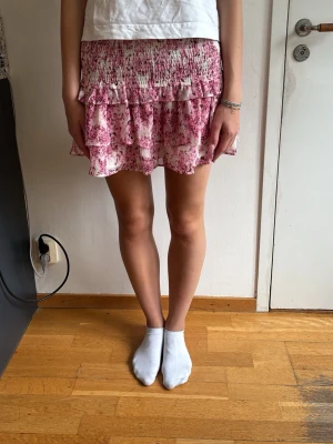 Rosa blommig volangkjol - Söt kortkjol i rosa och vitt med blommigt mönster och volangdetaljer. Kjolen är från Cos ursprungligen! Snygg att ha bara med en vit topp🩵