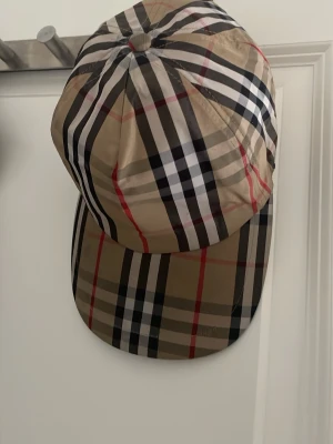 Burberry klassisk rutig keps - Säljer en snygg keps från Burberry med det ikoniska rutmönstret i beige, svart, vitt och rött. Kepsen har justerbar snapback och en patch bak med texten 'Burberry London England'. Perfekt accessoar för en trendig streetwear-look.
