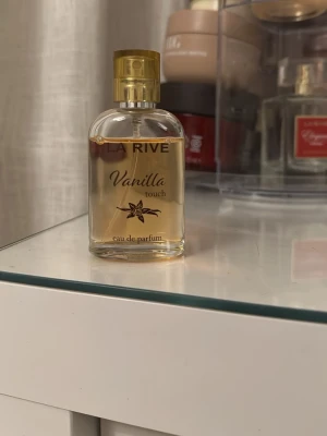 La Rive Vanilla Touch parfym - La Rive Vanilla Touch eau de parfum med en söt och varm vaniljdoft. Flaskan är genomskinlig med guldfärgat lock och text, och har en enkel, rundad form. Perfekt för dig som gillar mjuka och krämiga doftnoter.