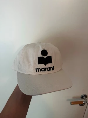 Vit keps från Isabel Marant - Snygg vit keps från Isabel Marant med svart broderad logga och text framtill. Klassisk form med böjd skärm och justerbar baksida. Tillverkad i ett tåligt tyg som passar perfekt till streetwear eller casual outfits.