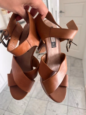 Bruna chunky sandaler med klack - Snygga bruna sandaler från Tamaris med breda korslagda remmar och kraftig klack. Sandalerna har justerbart spänne vid ankeln och en stadig platåsula. Tillverkade i läderimitation med en trendig retrovibe.