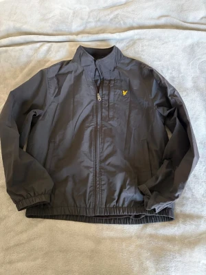 Svart vindjacka från Lyle & Scott - Snygg svart vindjacka från Lyle & Scott med dragkedja framtill och två sidofickor. Jackan har resår vid ärmslut och nederkant samt det klassiska gula logomärket på bröstet. Tillverkad i lätt och tåligt material, perfekt för blåsiga dagar.