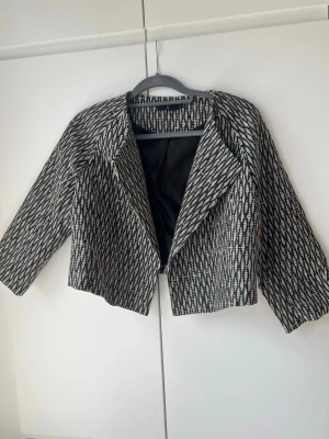 Kort mönstrad kavaj från Åhléns - Säljer en midjekort, svartvit kavaj från Åhléns i storlek 38. Kavajen har ett grafiskt mönster, breda slag och trekvartsärmar. Materialet är vävt och fodret är svart. Perfekt för dig som vill ha en unik och trendig look.