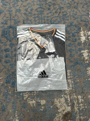 Svart Tyskland Adidas matchtröja - Snygg svart matchtröja från Adidas med Tysklands landslagsmärke på bröstet. Tröjan har vita ränder på axlarna, orange och röd kant vid hals och ärmslut samt Adidas-logga. Tillverkad i lätt och ventilerande material, perfekt för fotboll eller träning.