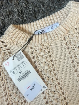 Beige stickad topp från Zara, ny med PRISLAPP!! - Snygg beige stickad topp från Zara med korta ärmar och rund hals. Tröjan har ett coolt hålmönster i ränder framtill och ribbade kanter. Perfekt till jeans eller kjol för en trendig look.
