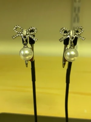 Söta örhängen med pärlor och rosett - Ett par silverfärgade örhängen med gnistrande rosett upptill och hängande vita pärlor. Rosetterna är dekorerade med små stenar som ger extra glans. Perfekt accessoar för att lyfta din stil med en söt och elegant touch.