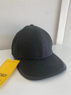 Svart keps från Fendi med FF-mönster - Snygg svart keps från Fendi med diskret FF-logga invävd i tyget. Klassisk böjd skärm med läderdetalj runt kanten och justerbar passform. Perfekt accessoar för dig som gillar exklusiv och stilren streetwear.