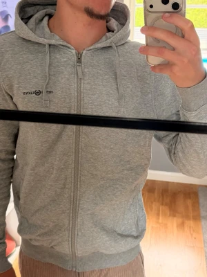 Grå zip hoodie från Henri Lloyd - Säljer en grå hoodie från Henri Lloyd med dragkedja och huva. Tröjan har dragsnören i huvan och ett diskret broderat Henri Lloyd-märke på bröstet. 