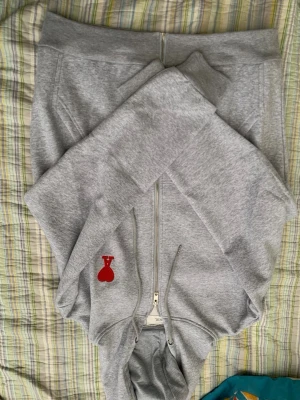 Grå zip hoodie från Ami Paris - Säljer en grå hoodie med dragkedja från Ami Paris. Hoodien har en huva med dragsko och en röd Ami de Coeur-logga broderad på bröstet. Tillverkad i mjuk bomullsblandning och har två fickor framtill. Perfekt för en chill och stilren look.