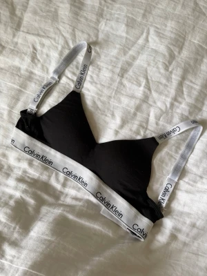 Svart Calvin Klein bralette - Snygg svart bralette från Calvin Klein med vita elastiska band och logotypdetaljer. Mjuk och bekväm modell utan bygel, justerbara axelband och hakknäppning bak. Perfekt för dig som gillar enkel och stilren design.