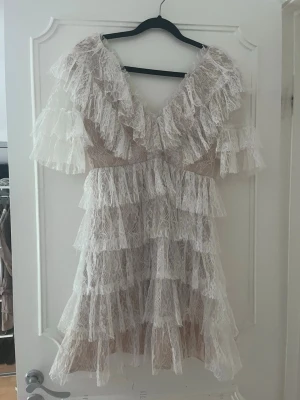 By malina klänning  - Supersöt kort klänning i vit spets med flera lager volanger och v-ringning både fram och bak. Klänningen har korta ärmar och ett romantiskt uttryck med transparent spets över beige foder. Perfekt för dig som vill sticka ut med en feminin look.