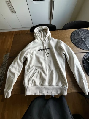 Vit hoodie från Better Bodies - Säljer en vit hoodie från Better Bodies med svart logga på bröstet. Hoodien har huva med dragsko och en stor magficka framtill. Tillverkad i mjukt material och har ribbade muddar vid ärmslut och nederkant. Perfekt för en avslappnad stil.