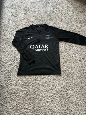 Svart PSG träningströja från Nike - Snygg svart långärmad träningströja från PSG med halv dragkedja framtill. Tröjan har vita logotyper från Nike och PSG samt Qatar Airways tryckt på bröstet. Ärmarna har diskret mönster och materialet är lätt och andas, perfekt för fotbollsträning.