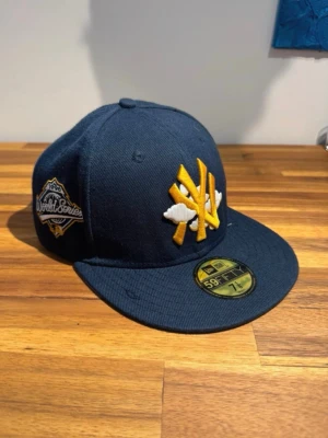 New Era Yankees 59FIFTY keps blå/gul - Snygg mörkblå New Era 59FIFTY keps med gul och vit Yankees-logga framtill och World Series-märke på sidan. Platt skärm med grå undersida och broderad detalj under skärmen. Klassisk streetwear-stil, perfekt för dig som gillar baseball och sportmode.