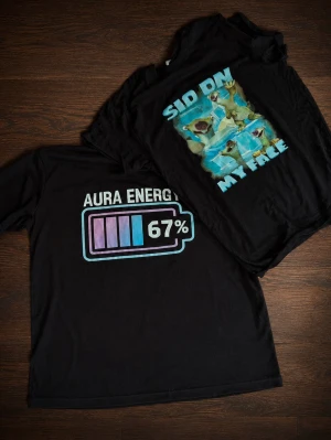 Svarta t-shirts med tryck - Två svarta t-shirts med coola tryck. Ena har texten 'AURA ENERGY 67%' och en batterisymbol i lila och blått. Den andra har texten 'SIT ON MY FACE' och ett färgglatt motiv med sengångare. Perfekta för dig som gillar statement-plagg.