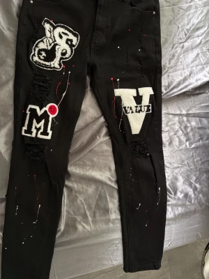 Svarta jeans med patches och tryck - Unika svarta jeans med stora patches och tryck i vitt och rött. Dekorerade med bokstäverna S, M och V samt ordet VALUE. Jeansen har distressed detaljer och färgstänk för en streetwear-look. Perfekta för dig som vill sticka ut med din stil.