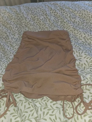 Beige ribbad kortkjol med snörning - Supersnygg beige kortkjol i ribbat material med snörning på båda sidorna som gör att du kan bestömma längden, Kjolen har en tight passform och rynkade detaljer som ger en trendig look. Perfekt att matcha med en crop top eller oversized hoodie.