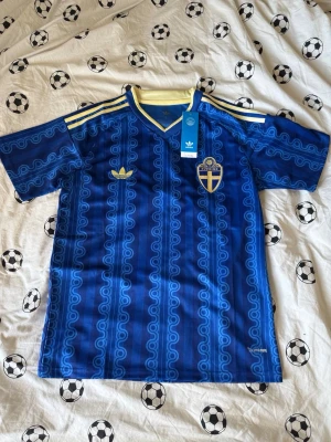 Blå Sverige fotbollströja Adidas - Säljer en blå Sverige fotbollströja från Adidas med retroinspirerat mönster, gula detaljer och broderad landslagssköld. Tröjan har korta ärmar, V-ringning och klassiska Adidas-ränder på axlarna. Perfekt för match eller träning.