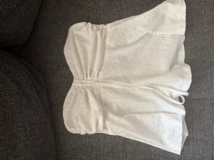 Vit axelbandslös topp med broderi - Superfin vit axelbandslös topp med broderade hål och resår. Lätt och luftig. Perfekt för varma dagar och enkel att styla.