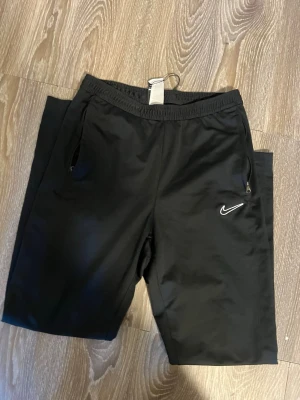 Svarta shorts från Nike med dragkedja - Svarta sportiga shorts från Nike med elastisk midja och dragkedjor på fickorna. Klassisk Nike-logga på vänster ben. Perfekta för träning eller chill. Tillverkade i ett lätt syntetmaterial som andas.Skriv för fler frågor och bilder