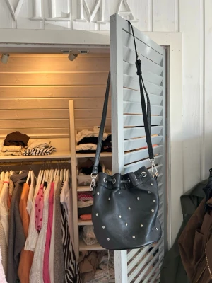 Svart bucket bag med nitar - Snygg svart axelväska i bucket-modell med silverfärgade nitar och dragskostängning. Väskan har justerbar axelrem och är gjord i ett slätt konstläder. Perfekt storlek för det viktigaste och ger en cool touch till din outfit.