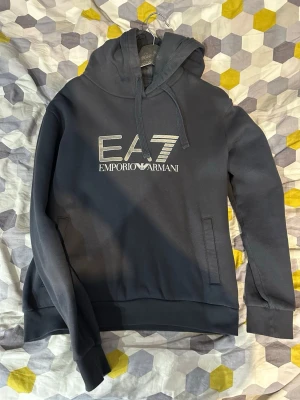 Mörkblå EA7 hoodie med huva - Snygg mörkblå hoodie från Emporio Armani EA7 med stor logga framtill. Tröjan har huva med dragsko och praktiska fickor på magen. Tillverkad i mjukt material som känns skönt mot huden. Perfekt för en avslappnad och trendig look.