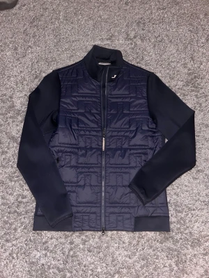 Marinblå quiltad jacka J.Lindeberg - Snygg marinblå quiltad jacka från J.Lindeberg med dragkedja framtill och hög krage. Jackan har släta ärmar och ett diskret logotryck på kragen. Perfekt för dig som vill ha en stilren och sportig look.