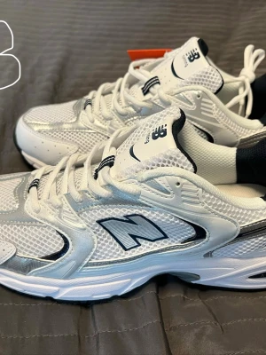 New Balance 530 vita sneakers - Säljer ett par fräscha New Balance 530 sneakers i vitt och silver med mesh och syntetdetaljer. Klassisk chunky sula och svart New Balance-logga på sidan. Perfekta för dig som gillar retrostil och bekväma skor.