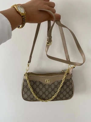 Beige axelväska i konstläder - Stilren beige axelväska i konstläder med enkel, rektangulär form och justerbar axelrem. Väskan har guldfärgade metalldetaljer och en slät yta som ger en modern look. Perfekt för dig som gillar minimalistisk stil. Helt ny luktar lite PU leder men försvinner efter att du har använd det några gånger. Skriv gärna till mig innan köp