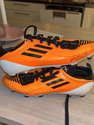 Adidas F50 orange fotbollsskor - Säljer ett par grymma Adidas F50 fotbollsskor i orange med svarta detaljer och svarta snören. Skorna har klassiska tre svarta ränder på sidan, låg profil och dobbar för spel på gräs. Materialet är syntetiskt och designen är riktigt sportig och snabb. Pris kan diskuteras
