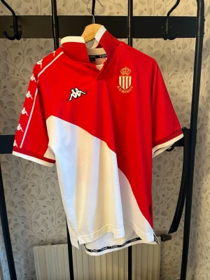 AS Monaco matchtröja från Kappa - Säljer en AS Monaco matchtröja från Kappa i rött och vitt med klubbmärke på bröstet och Kappa-loggor längs ärmarna. Tröjan har en modern krage och är tillverkad i ett lätt och ventilerande syntetmaterial, perfekt för fotboll eller träning.