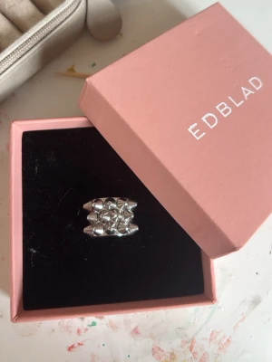 Silverfärgad ring från Edblad - Stilren silverfärgad ring från Edblad med flera små, blanka pärlliknande detaljer i ett rutmönster. Ringen har en bred design och är tillverkad i metall med en modern och trendig look. Perfekt accessoar för dig som gillar unika smycken.