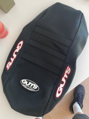 Guts säte - Säljer ett svart sadelöverdrag från Guts Factory med texturerad yta och logga i vitt och rött. Överdraget har ribbor för extra grepp och är gjort i slitstarkt material, perfekt för motocross eller offroad.