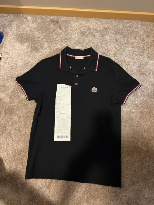 Svart Moncler pikétröja med röd/vit kant - Klassisk svart pikétröja från Moncler med röd och vit kant på krage och ärmslut. Tröjan har Moncler-logga på bröstet och knäppning framtill. Perfekt för en stilren och sportig look. Köpt på nk och har kvar kvittot. 100% äkta