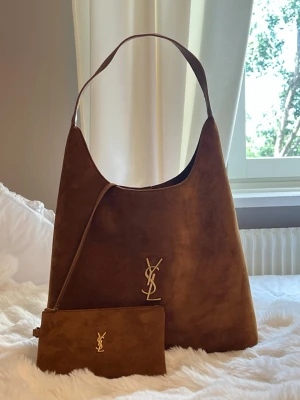 Brun mocka handväska från YSL - Snygg brun handväska i mocka från Yves Saint Laurent med guldigt YSL-emblem framtill. Väskan har en mjuk, slouchy form och bred axelrem. Tillhörande liten clutch i samma material och färg med matchande logga. Perfekt accessoar för en trendig look. Köpt i Dubai 