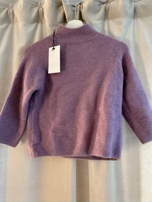 Ljuslila stickad tröja från Zara - Mysig stickad tröja från Zara i en ljuslila nyans. Tröjan har hög krage, raka ärmar och ribbstickade detaljer vid hals, ärmslut och nederkant. Perfekt för lager-på-lager och enkel att matcha med jeans eller kjol.