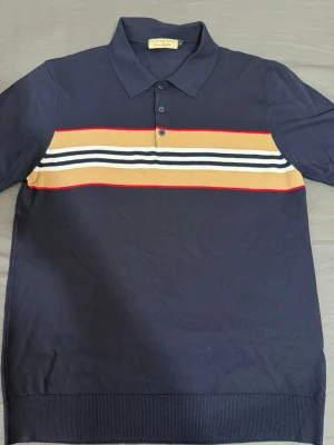 Mörkblå Burberry piké med ränder - Snygg mörkblå pikétröja från Burberry med klassisk krage och knappar. Tröjan har horisontella ränder i beige, vit, svart och röd över bröstet. Perfekt för en stilren och trendig look. S\M