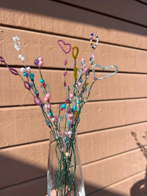 Handgjorda pärlblommor på ståltråd - Unika handgjorda dekorationer av pärlor i olika färger och former, formade som blommor och hjärtan på gröna ståltrådar. Perfekt kreativ accessoar till ditt rum. Pärlorna är i rosa, blått, vitt, gult och lila och ger en lekfull och personlig touch. (Vasen ingår ej) 