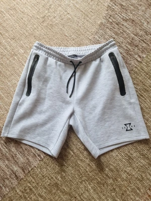 Grå Contra Shorts  - Snygga ljusgrå shorts från Contra med svarta dragkedjor på fickorna och snörning i midjan. Perfekta för sommaren och har en modern, sportig look. Tillverkade i mjukt material som känns skönt mot huden. Contra-logga tryckt på ena benet.
