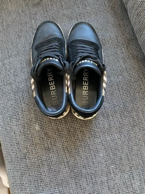Svarta Burberry sneakers med rutmönster - Snygga svarta sneakers från Burberry med klassiskt beige, svart och rött rutmönster på sidorna. Skorna har vita sulor, svarta skosnören och är tillverkade i läder och textil. Perfekta för dig som vill ha en stilren och exklusiv look.