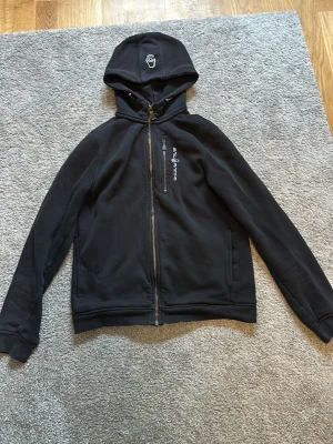 Svart hoodie från Sail Racing - Svart hoodie från Sail Racing med dragkedja framtill och huva. Har en bröstficka med dragkedja och Sail Racing-logga på bröstet samt tryck på huvan. Mjuk insida och ribbade muddar. Perfekt för en chill och sportig stil.
