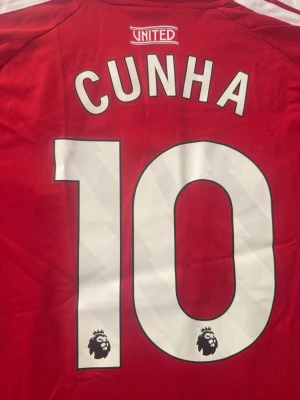 Röd Manchester United matchtröja - Snygg röd Manchester United matchtröja med CUNHA och nummer 10 på ryggen. Premier League-logga och 'No room for racism'-tryck på ärmen. Klassisk design med vita detaljer och ränder på axlarna. Perfekt för fotbollsfans!
